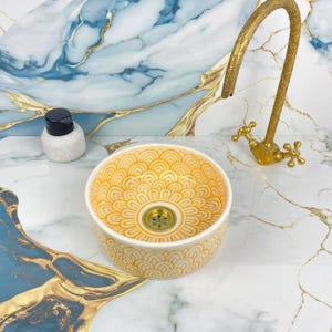 Peut inclure: Un évier rond en céramique avec un motif floral jaune et blanc. Un robinet et un drain dorés sont visibles. Une petite bouteille blanche et noire est sur le comptoir. L'évier est sur un comptoir en marbre.