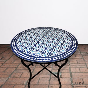 Może przedstawiać: Okrągły stół z blatem z mozaiki z niebieskich i białych płytek oraz czarną metalową ramą. Stół stoi na patio z cegły.