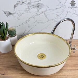 Puede incluir: Un lavabo de cerámica blanca con un borde dorado y un desagüe de latón. El lavabo está sobre una encimera de madera con un grifo cromado.
