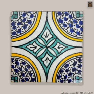 Może przedstawiać: Kwadratowa płytka ceramiczna z geometrycznym wzorem w kolorze niebieskim, zielonym, żółtym i białym. Płytka ma centralny kształt rombu z czterema mniejszymi rombami w każdym rogu. Płytka jest ozdobiona motywem kwiatowym.