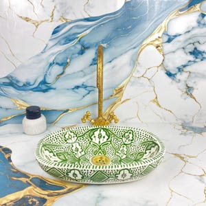Peut inclure: Un lavabo ovale en céramique avec un motif floral vert et blanc. Un robinet et un drain dorés sont installés. Un petit récipient blanc avec un couvercle bleu foncé est sur le comptoir. Le fond est un motif de marbre avec des accents bleus et dorés.