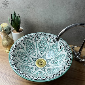 Może przedstawiać: Biała ceramiczna umywalka z zielonym i czarnym geometrycznym wzorem. Umywalka ma odpływ z mosiądzu i jest zamontowana na drewnianym blacie.