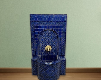 May include: A blue mosaic wall fountain with a gold-colored faucet. The fountain has a rectangular back and a curved basin. The mosaic tiles are in shades of blue. The fountain is mounted on a wall with a light green background.