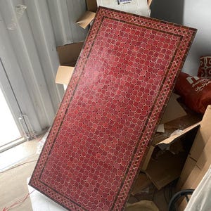 Könnte beinhalten: Rechteckiger Tisch mit roter Mosaikfliesenoberfläche und geometrischem Muster. Dunkelbrauner Rand. Umgeben von Kartons und anderen Gegenständen. Abmessungen nicht sichtbar.