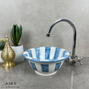 Könnte beinhalten: Ein weißes Keramikwaschbecken mit blauen und weißen Streifen. Das Waschbecken ist rund und hat eine leichte Wölbung. Das Waschbecken steht auf einer grauen Arbeitsplatte mit einem verchromten Wasserhahn.