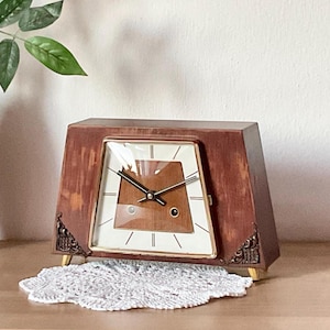 Puede incluir: Un reloj de sobremesa trapezoidal de madera con acabado marrón y detalles metálicos dorados en las esquinas inferiores. La esfera presenta un fondo blanco con finas marcas horarias negras, manecillas negras y una incrustación geométrica.