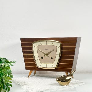 May include: A trapezoidal mantel clock with a dark wood grain finish and horizontal gold-toned inlay stripes. The clock face is cream-colored with gold hands and the text "flamla" printed in the center, encased in a gold-toned frame.