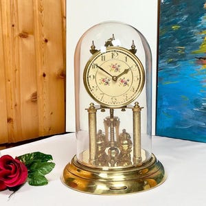 May include: A vintage brass clock with a floral-decorated face, encased in a glass dome. The clock features a pendulum and is set on a round brass base. The clock's face has black numerals and hands, with floral accents.