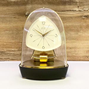 May include: A gold-toned, triangular-shaped clock with a white face and black hands. The clock is encased in a clear, dome-shaped glass cover and sits on a black base.