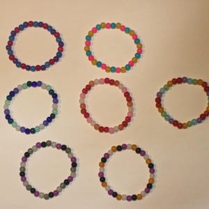May include: Seven beaded bracelets in various colors, including blue, pink, green, and yellow. The bracelets are made of small, round beads and are arranged in a circular shape. The bracelets are on a white surface.