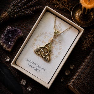 May include: A gold-coloured Sacred Trinity necklace in a gift box. The pendant has a Celtic knot design. The box contains a card with the words "Magickly Woven" and "Sacred Trinity Necklace". The background includes a lit candle, crystals, and a book.