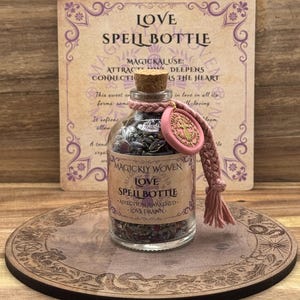 May include: A glass spell bottle labelled "LOVE SPELL BOTTLE" filled with dried herbs and crystals. The bottle has a cork stopper and a pink tag. A decorative wooden coaster and a card with text about love are in the background.