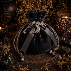 May include: A dark grey velvet drawstring pouch featuring a silver-toned metal charm set consisting of a skeleton key, a crescent moon, and a star. The pouch is cinched with a matching grey satin ribbon and braided cords ending in small silver beads.