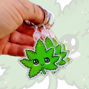 May include: Three green glitter acrylic keychains shaped like marijuana leaves with a cartoon face. Each keychain has a silver metal ring.