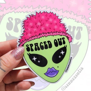 May include: A sticker featuring a green alien face with large black eyes, star-shaped pupils, and purple lips. The alien has pink, fluffy hair and the words "SPACED OUT" in black. The sticker is held by a hand with a glittery nail.