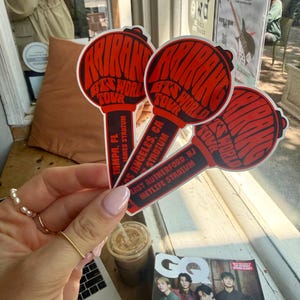 May include: Three red and white stickers with the text "BTS WORLD TOUR" in a stylized font. The stickers are shaped like microphones and list stadium locations: Tampa, FL, Los Angeles, CA, and East Rutherford, NJ. A magazine with a group of men is visible.