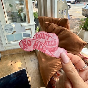 May include: A pink sticker in the shape of a cowboy hat with the words "DON'T NEED NO MESS" in a retro font. The sticker is held up by a person's hand with a ring on the finger. A laptop and a drink are on the table.