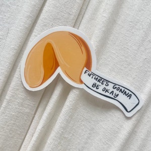 May include: A sticker featuring a fortune cookie design. The cookie is a warm orange color with a white paper fortune that reads "Future's Gonna Be Okay" in black text. The sticker is on a white fabric background.