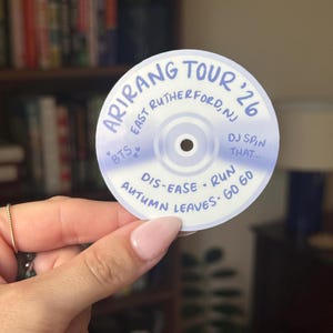 May include: A circular sticker designed to look like a compact disc with a light blue and white gradient. It features the text Arirang Tour '26, East Rutherford, NJ, BTS, DJ Spin That, Dis-ease, Run, Autumn Leaves, and Go Go in blue ink.