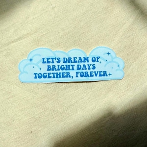 May include: A light blue sticker in the shape of a cloud with the text "LET'S DREAM OF BRIGHT DAYS TOGETHER, FOREVER+" in blue. The sticker has small blue crosses and stars around the cloud.