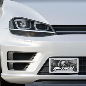 May include: Close-up of a white car's front end, showcasing a license plate with a skull design and the text "F-THIS!". The car's headlights and bumper are also visible.
