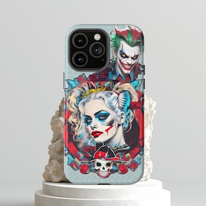 May include: A phone case featuring a colorful illustration of Harley Quinn and the Joker. The design includes a portrait of Harley Quinn with red and black makeup, and the Joker with green hair and a sinister smile. The case has a textured, light-colored background.