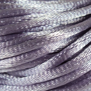 Puede incluir: Primer plano de una bobina de hilo de bordar de color lavanda. El hilo está enrollado apretado y tiene un brillo sutil.