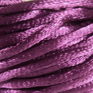 Puede incluir: Primer plano de un carrete de cordón de satén morado oscuro. El cordón es suave y brillante, con un brillo sutil.
