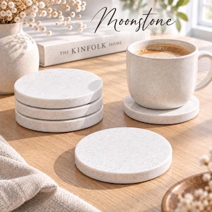 Pode incluir: Um conjunto de bases para copos redondas em pedra branca salpicada com acabamento liso. Uma base está colocada sob uma caneca de cerâmica branca salpicada a condizer com café, enquanto uma pilha de três bases adicionais repousa por perto.