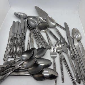 May include: A collection of assorted stainless steel flatware including various spoons, forks, and knives. The set features pieces with decorative embossed patterns on the handles, alongside plain utensils, a slotted spoon, and a cake server.