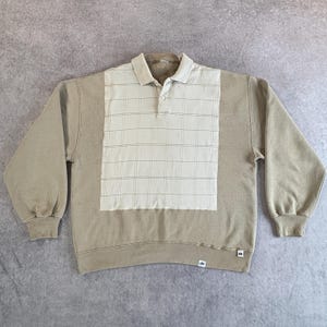 May include: A tan-colored sweatshirt with a collared polo-style neckline. The front panel features a light beige plaid pattern. The sleeves and lower body are a solid tan color. The sweatshirt has a relaxed fit and long sleeves.