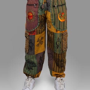 May include: Multi-coloured patchwork trousers with embroidered sun, moon, and other patterns. The trousers combine green, yellow, and purple panels. They have pockets and zips, and are worn with white trainers.