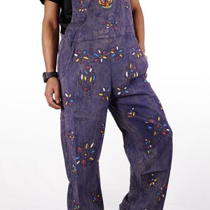 May include: Blue denim overalls with colorful floral embroidery and a mushroom design. The overalls have a relaxed fit and are perfect for a casual, bohemian look.