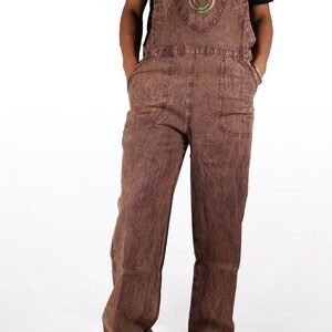 May include: Brown denim overalls with a green and red spiral design on the chest pocket. The overalls have adjustable straps and two front pockets.