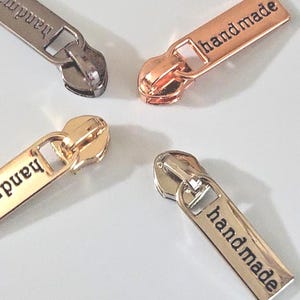 May include: Four metal zipper pulls in silver, rose gold, and gold tones, each featuring a rectangular tab engraved with the word handmade. The pulls are arranged in a circular pattern, and the text JessmargaShopBags&More appears at the bottom.