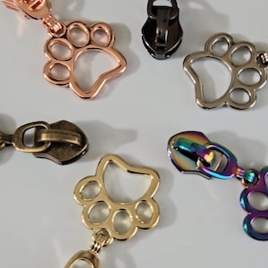 May include: A collection of six metal zipper pulls in various finishes including rose gold, black, silver, antique brass, gold, and iridescent rainbow. Each pull features a cutout paw print design. The text JessmargaShopBags&More is visible.