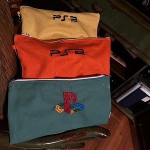 May include: Three canvas zipper pouches in yellow, orange, and teal. The yellow and orange pouches have the embroidered letters "PS3". The teal pouch features a colorful embroidered PlayStation logo. Each pouch has a white zipper.