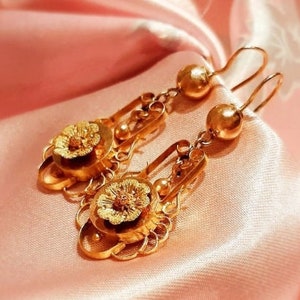 May include: A pair of gold dangle earrings featuring a floral design. The earrings have a round, gold bead at the top and a smaller, round, gold bead at the bottom. The floral design is intricate and detailed.