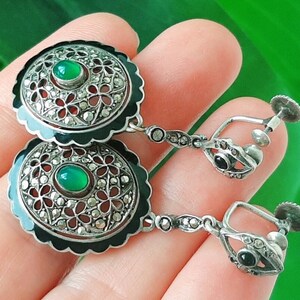 May include: A pair of silver clip-on earrings with black enamel and green gemstones. The earrings feature a delicate filigree design and are accented with marcasite stones.