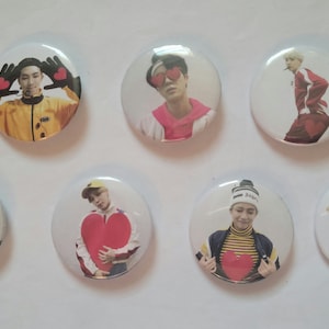 Op de afbeelding: Zeven ronde knoopsluitingen met verschillende afbeeldingen van een K-pop boyband. Elke knoopsluiting toont een ander bandlid met een andere uitdrukking en pose. De knoopsluitingen zijn allemaal wit met een zilveren rand.