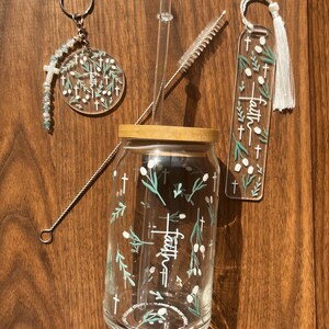 May include: A clear glass tumbler with a bamboo lid, a keychain, and a bookmark, all featuring the word "faith" and cross designs. The tumbler includes a clear straw and a cleaning brush. The items have a floral and cross theme.