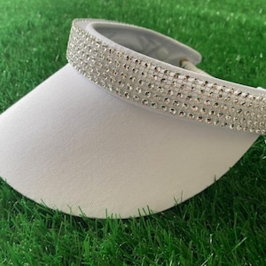 May include: A white visor with a curved brim and a band of sparkling rhinestones. The visor is set against a backdrop of green grass, suggesting outdoor wear for sun protection.