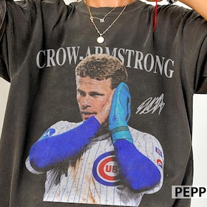 Pode incluir: Uma t-shirt cinzento-carvão de manga curta com uma impressão gráfica de um jogador de basebol com camisola às riscas, mangas azuis e luva azul. O texto CROW-ARMSTRONG está impresso acima do jogador, com uma assinatura à direita.