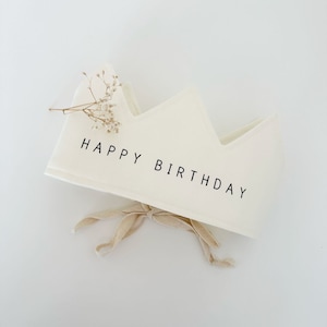 May include: White fabric crown with a beige ribbon and the text "HAPPY BIRTHDAY" printed in black.