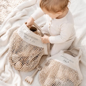 Puede incluir: Dos bolsas de malla beige con partes superiores de tela s&oacute;lida que presentan el texto SANDSPIELZEUG y nombres personalizados. Las bolsas contienen varios art&iacute;culos de color claro y un peque&ntilde;o rastrillo de pl&aacute;stico junto a la bolsa.