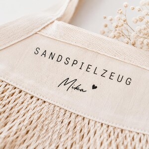 Puede incluir: Una bolsa de red de algod&oacute;n de color beige con un panel de tela s&oacute;lida en la parte superior. El panel tiene impreso el texto negro SANDSPIELZEUG y el nombre Mika seguido de un peque&ntilde;o s&iacute;mbolo de coraz&oacute;n. La bolsa tiene asas tejidas.