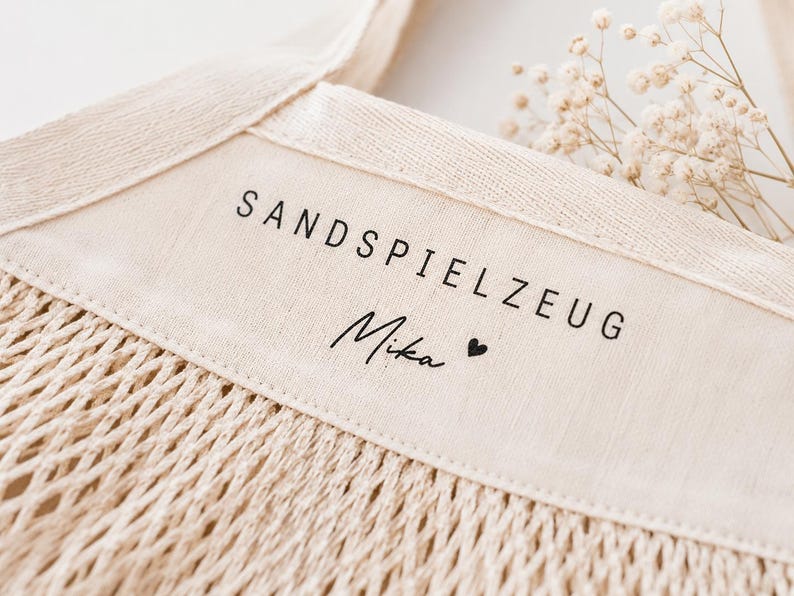 Puede incluir: Una bolsa de red de algod&oacute;n de color beige con un panel de tela s&oacute;lida en la parte superior. El panel tiene impreso el texto negro SANDSPIELZEUG y el nombre Mika seguido de un peque&ntilde;o s&iacute;mbolo de coraz&oacute;n. La bolsa tiene asas tejidas.