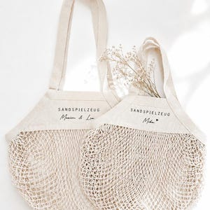 Puede incluir: Dos bolsas de malla de algod&oacute;n en color blanco roto con paneles superiores de tela s&oacute;lida. Cada bolsa presenta el texto impreso en negro SANDSPIELZEUG. Una bolsa est&aacute; personalizada con Maxim & Lea, y la otra con Mika y un peque&ntilde;o coraz&oacute;n.