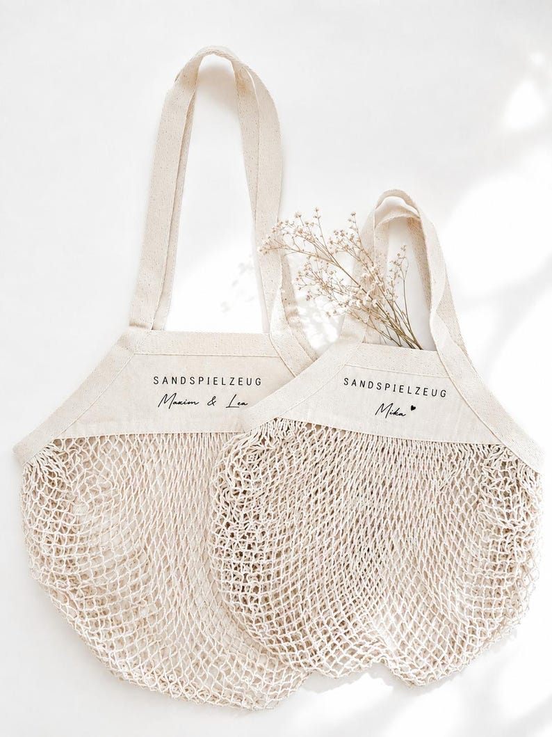 Puede incluir: Dos bolsas de malla de algod&oacute;n en color blanco roto con paneles superiores de tela s&oacute;lida. Cada bolsa presenta el texto impreso en negro SANDSPIELZEUG. Una bolsa est&aacute; personalizada con Maxim & Lea, y la otra con Mika y un peque&ntilde;o coraz&oacute;n.