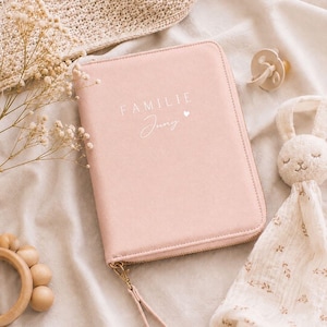 May include: A rectangular, light pink zippered organizer case with a wrist strap. The front cover features the white printed text "FAMILIE" above the script text "Jung" and a small heart icon. The case has a smooth, solid-colored exterior finish.